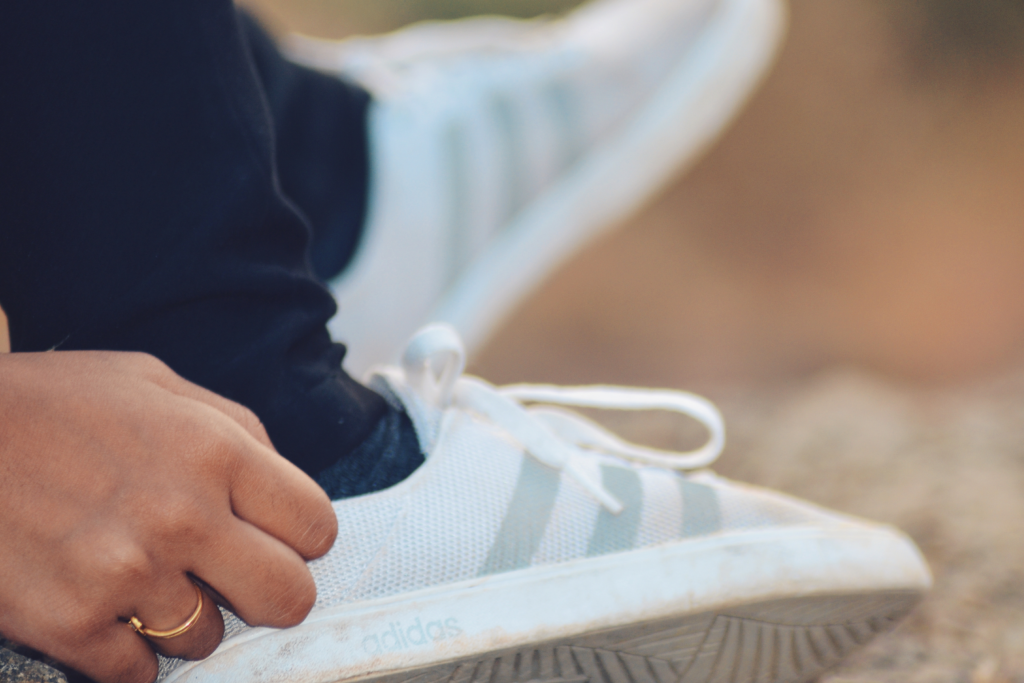 Hier sind zwei weiße Adidas Schuhe zu sehen, die von einer Person getragen werden, welche bei einem schlichten Hintergrund präsentiert werden. Die Farbe der Hose ist ein dunkles Blau.