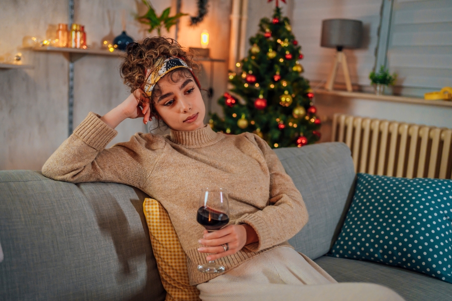 Frau sitzt auf dem Sofa, denkend mit einem Glas Rotwein in der Hand, ein Christbaum ist im Hintergrund. Philosophie am Abend