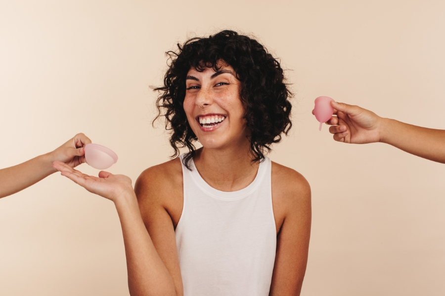 Frau lächelt und bekommt von zwei Seiten eine Menstruationstasse und eine Menstruationsscheibe gereicht. Sie selbst hält die Hand zur Menstruationsscheibe hin.