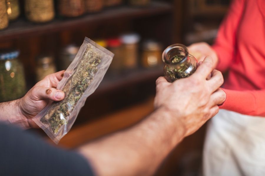 In einer Apotheke verkauft eine Person einer anderen eingeschweißte Hanfblüten, die eine Hand gibt der anderen ein Glas mit Blüten.